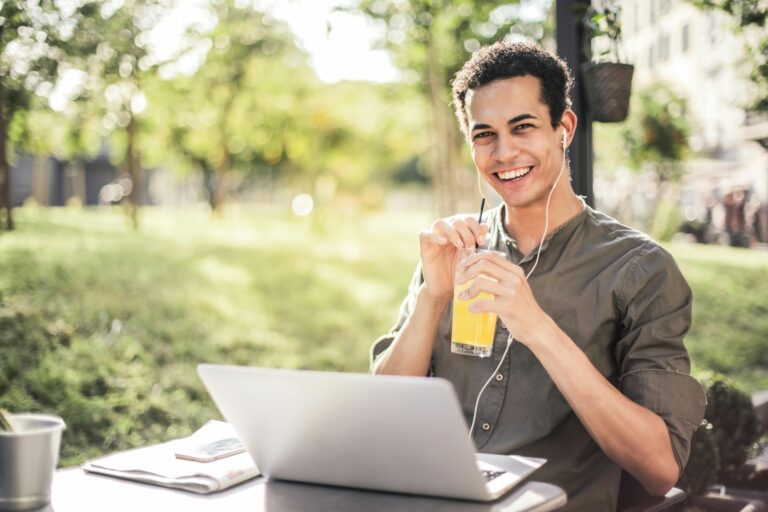 happy man wearing wires headphones standing at a desk working on a laptop and drinking a juice in nature