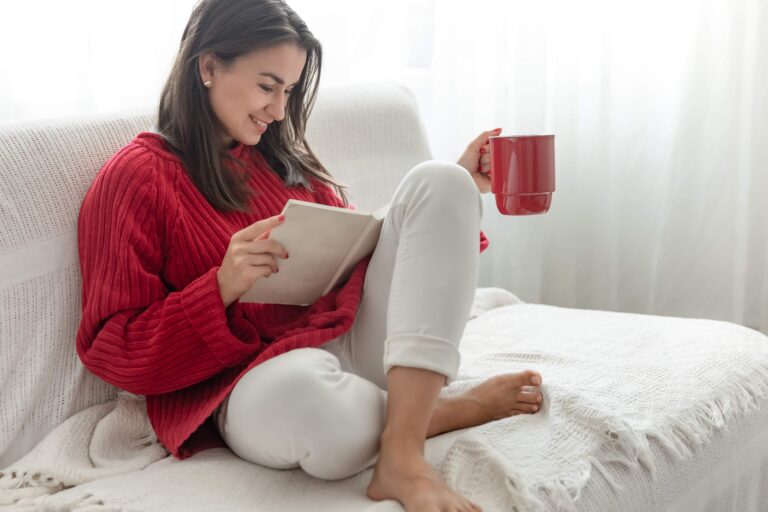 Young attractive woman in a cozy red sweater is reading a book while sitting at home on the sofa with a cup of drink.
